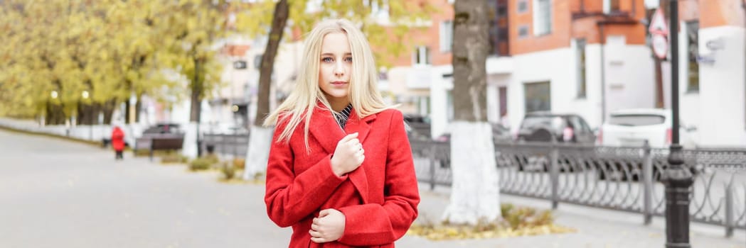 A young blonde woman walks around the autumn city in a red coat. The concept of urban style and lifestyle.