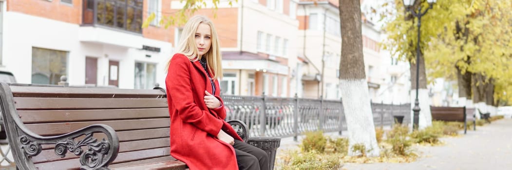 A young blonde woman walks around the autumn city in a red coat. The concept of urban style and lifestyle.