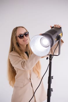 Professional gaffer working, adjusts constant light. Woman blonde smiling in sunglasses, beige suit on white isolated background in photo studio. Vertical