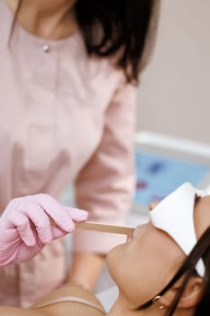 Close-up of beautician holds a wooden spatula and applies a transparent gel with anesthetic to the woman's face and prepares the skin for the laser hair removal procedure.