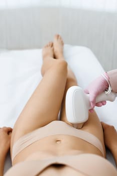 Anonymous beautician in gloves using laser hair removal apparatus on bikini zone of female client during beauty laser skin care procedure on stomach in modern salon. Laser epilation at beauty clinic.