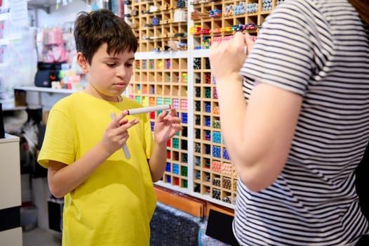 Charming serious teenage boy chooses watercolor markers in school stationery store. Inspired creative child asks sales manager for help, standing by a shelf with huge assortment of color felt-tip pens