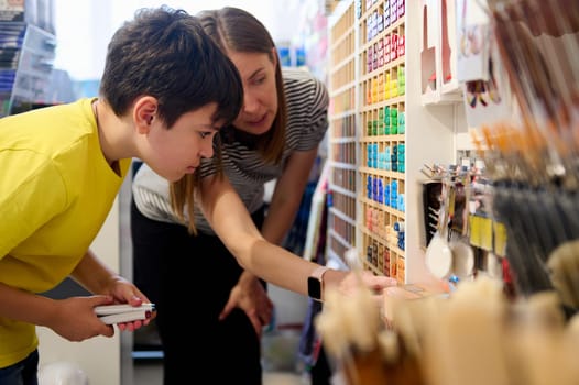 Adorable dark-haired teen schoolboy asking the advice of a sales manager in art shop, choosing felt-tip pens from the shelf with a large assortment of colorful drawing, painting and sketching tools