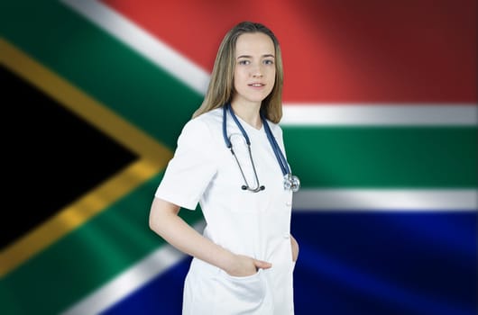 A young female doctor in a white coat and a stethoscope stands on the background of the flag of South Africa.