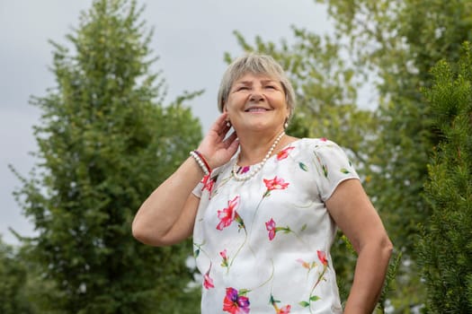 Real Smiling Senior 70 yo Woman Looks Away. Green Trees and Sky on Background. Portrait Of Happy Grandmother Outside, Enjoying Retirement Life. Healthy Old Mature Female, Lifestyle. Horizontal Plane.