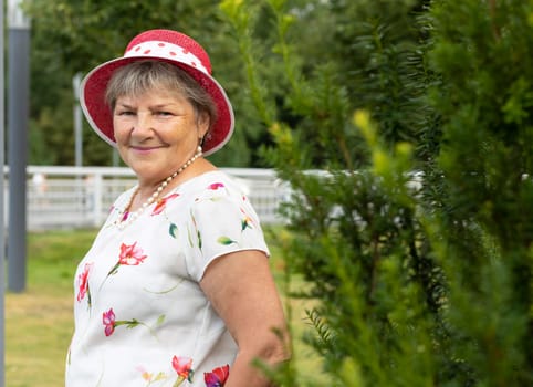 Real Smiling Senior Woman In Hat In Sunny Day Looks At Camera. Green Trees, Park on Background. Portrait Of Happy Elderly Granny, Enjoying Retirement Life, Vacation In Country House Horizontal Plane