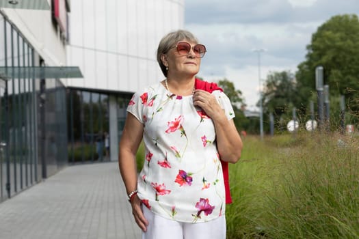 Real Modern Senior Woman In Sunglasses Walks Near Shopping Mall. Green Trees, Park on Background. Portrait Of Happy Elderly Female, Enjoying Retirement Life In City. Authentic Lifestyle Horizontal.