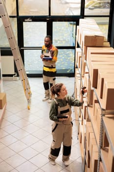 Employee scanning cardboard boxes using store scanner during warehouse inventory, typing products barcode on tablet computer. Supervisor in protective overall preparing clients orders in storehouse