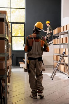 Warehouse supervisor checking carton boxes with products, typing goods details report on laptop computer during inventory in storage room. Employee with protective overall working in storehouse