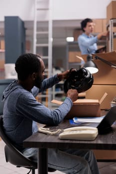 Employee looking at helmet checking product status before delivery package to customer, checking client shipping detalies on laptop computer. African american manager working in warehouse