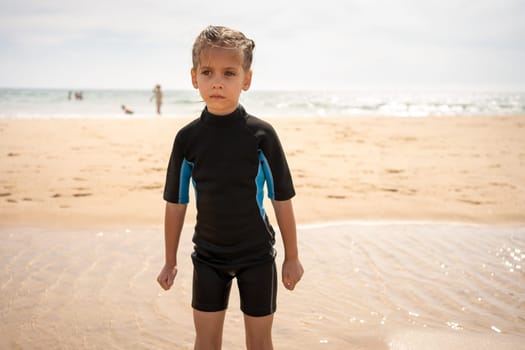 Little girl surfer in wetsuit standing ocean beach. Serious child after surfing on sandy beach