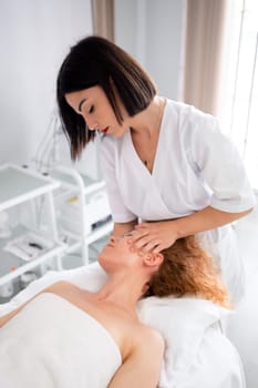 Massage therapist massaging head, shoulders and neck to woman in cosmetology clinic. Beautician making skincare and stimulation procedure. Spa treatment and massage in beauty salon. Vertical photo