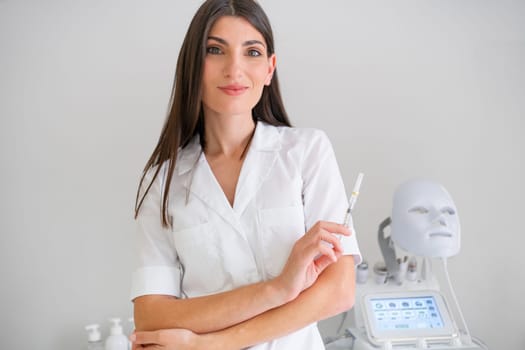 Woman cosmetologist standing hold injection syringe and looking at camera in beauty spa salon. Female dermatologist, skin therapist, beautician skincare professional in clean white coat uniform