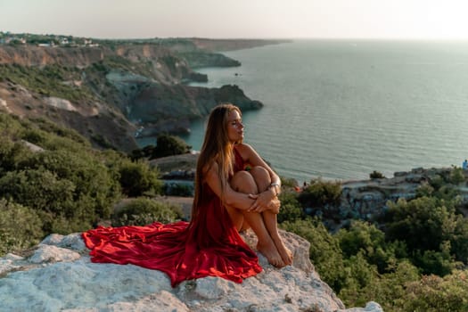 Woman sunset sea red dress, side view a happy beautiful sensual woman in a red long dress posing on a rock high above the sea on sunset