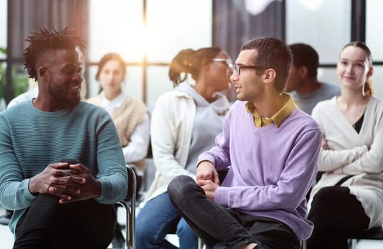 two men communicate while sitting on a chair waiting for a speaker in a conference room