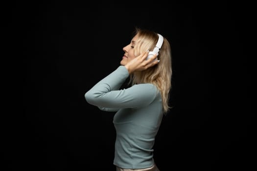 Profile portrait of charming blonde woman in massive white headphones listening to music on isolated black background