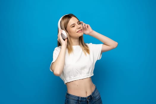 Beautiful blonde dancing young woman listening the music in white wireless headphone and holding mobile phone on blue background