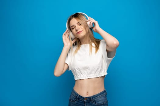 Optimistic blonde woman in a white t-shirt smiles pleasantly closes eyes rates song which she heard in white stereo headphones isolated over blue wall. Favorite song, enjoying cool playlist