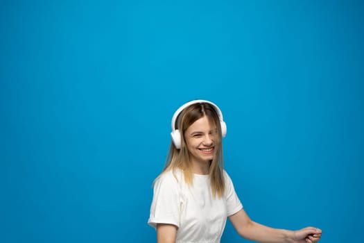 Happy blonde woman with a white headphones listening to music and dancing on a blue background. Woman moving to favorite song, enjoying cool playlist