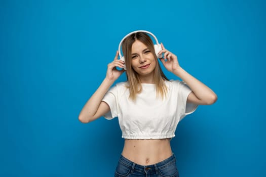 Energy girl with white headphones listening to music on blue background in studio. She wears white t-shirt and shorts