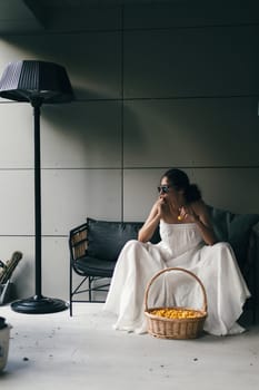 Portrait of a beautiful indian woman in white dress and sunglasses sitting on the sofa with a basket of yellow sweet cherry.