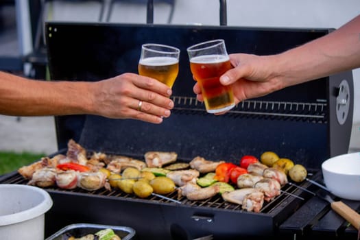 men hold beer in their hands on the background of the grill, vegetables and meat. Selective focus. Nature