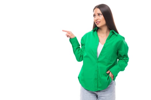 attractive young caucasian brunette lady with makeup dressed in an elegant green shirt points her finger on a white background with copy space.