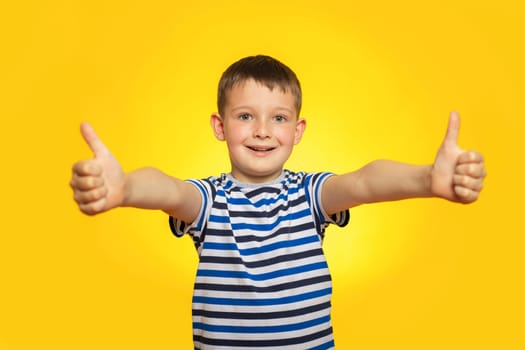 Portrait of happy boy in striped t-shirt showing thumbs up gesture on yellow background.