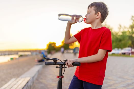 A boy drinks water from plastic bottle, standing with scooter.