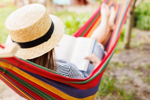 Young woman reading book while lying in comfortable hammock at green garden.