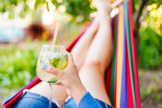 Young woman with a glass of cocktail resting lying in a comfortable hammock in a green garden.