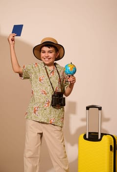 Happy smiling teenage boy wearing stylish summer casual clothes, holding globe and passport, smiling broadly looking at camera, standing with a yellow suitcase, isolated over white studio background
