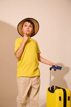 Handsome Caucasian teenage boy in stylish casual summer clothes, holding finger on his chin, thoughtfully looking aside a copy advertising space, standing with suitcase over white isolated background