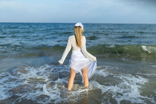 Beautiful girl in the sea. The woman enters the sea. Blue ocean. Girl in a white cape. Beautiful woman on the beach. Summer vacation at sea. The girl loves the sea.