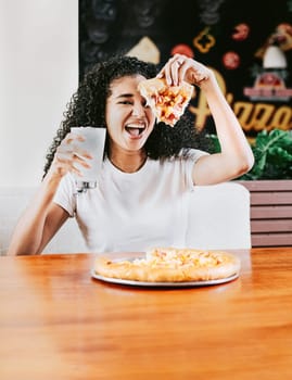 Happy afro hair woman showing slice of pizza in a restaurant. Smiling afro woman enjoying a pizza in a restaurant