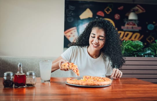 Happy afro hair woman eating pizza in a restaurant. Lifestyle of afro-haired woman enjoying a pizza in a restaurant
