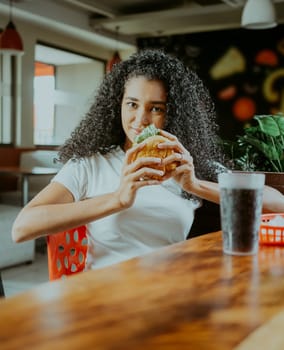 Beautiful latin girl enjoying a delicious hamburger in a restaurant. Portrait of afro girl holding a hamburger in a restaurant.