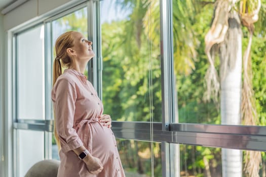 Portrait of young pregnant woman standing near window holding hands on her belly at modern home with copy space. Pregnancy, motherhood, expectation and tenderness concept.