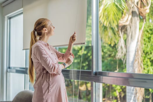 Portrait of young pregnant woman standing near window holding hands on her belly at modern home with copy space. Pregnancy, motherhood, expectation and tenderness concept.