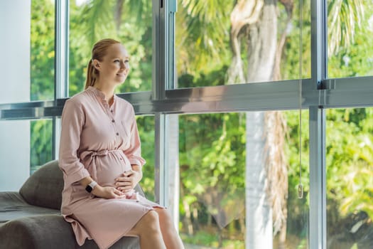 Portrait of young pregnant woman sitting near window holding hands on her belly at modern home with copy space. Pregnancy, motherhood, expectation and tenderness concept.