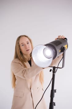 Professional female gaffer working, adjusts constant light. Woman blonde, beige suit on white isolated background in photo studio. Vertical, copy space