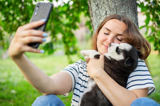 Young beautiful woman in a t-shirt and jeans with a husky puppy takes a photo or selfie on the phone while sitting in a park near a tree.