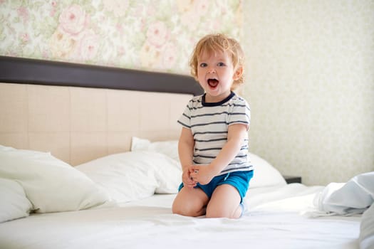 Cute laughing child boy sits in bed. Boy plays at home. Child in a sunny bedroom.