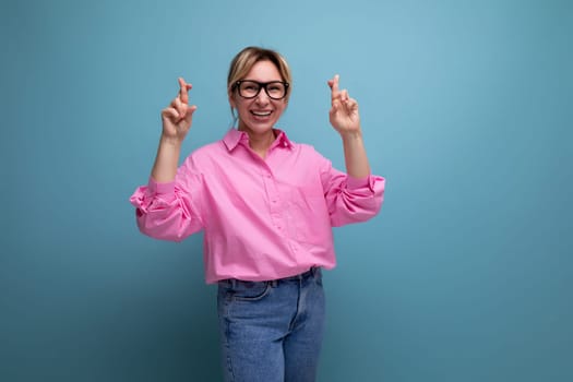 young caucasian blonde secretary woman with ponytail hairstyle dressed in pink shirt crossed her fingers on studio background with copy space.