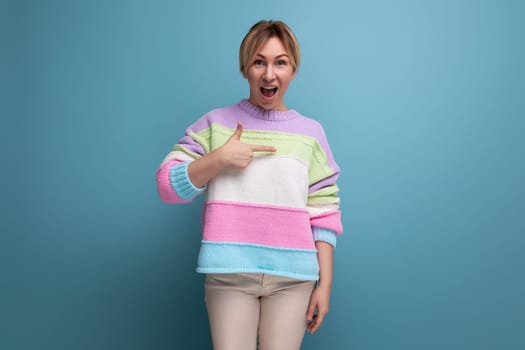 surprised blond young woman in casual outfit pointing at wall with finger on blue background with copy space.