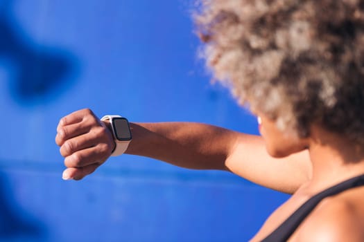 unrecognizable african american sportswoman looking at smart watch with blue background, concept of sports technology and active lifestyle, copy space for text