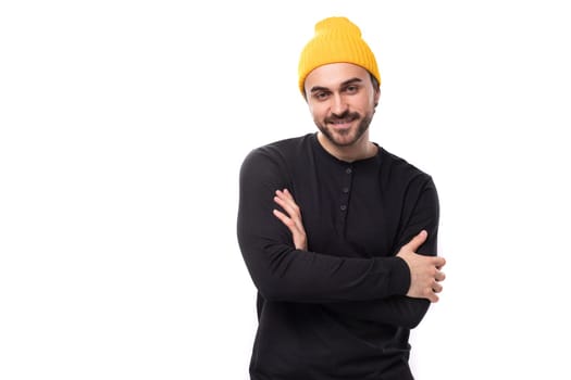 attractive young 30 year old guy with a beard dressed in a black jacket and a yellow hat looks at the camera.