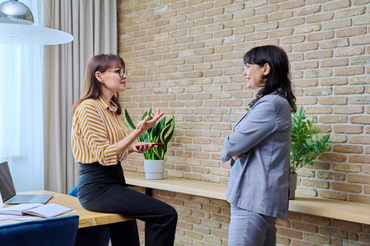 Two mature business women are talking in a modern office. Laughing communicating female colleague, 40s 50s age. Business, ceo, work, law, finance, mentoring, consulting, teamwork, job