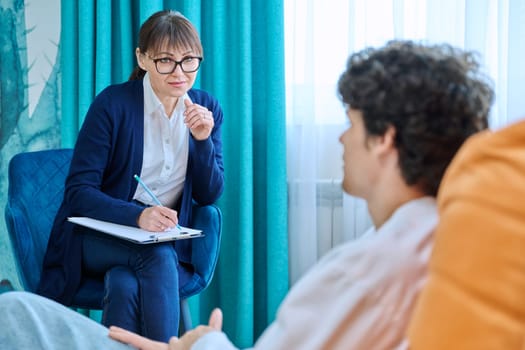 Middle aged confident friendly female psychologist talking to young male, individual therapy session, focus on therapist counselor social worker. Mental health support psychology psychotherapy concept