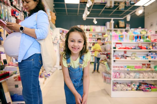 Caucasian adorable mischievous little child girl in casual denim, smiling broadly looking at camera, enjoying a shopping day with her mother in the school stationery shop, buying school supplies.